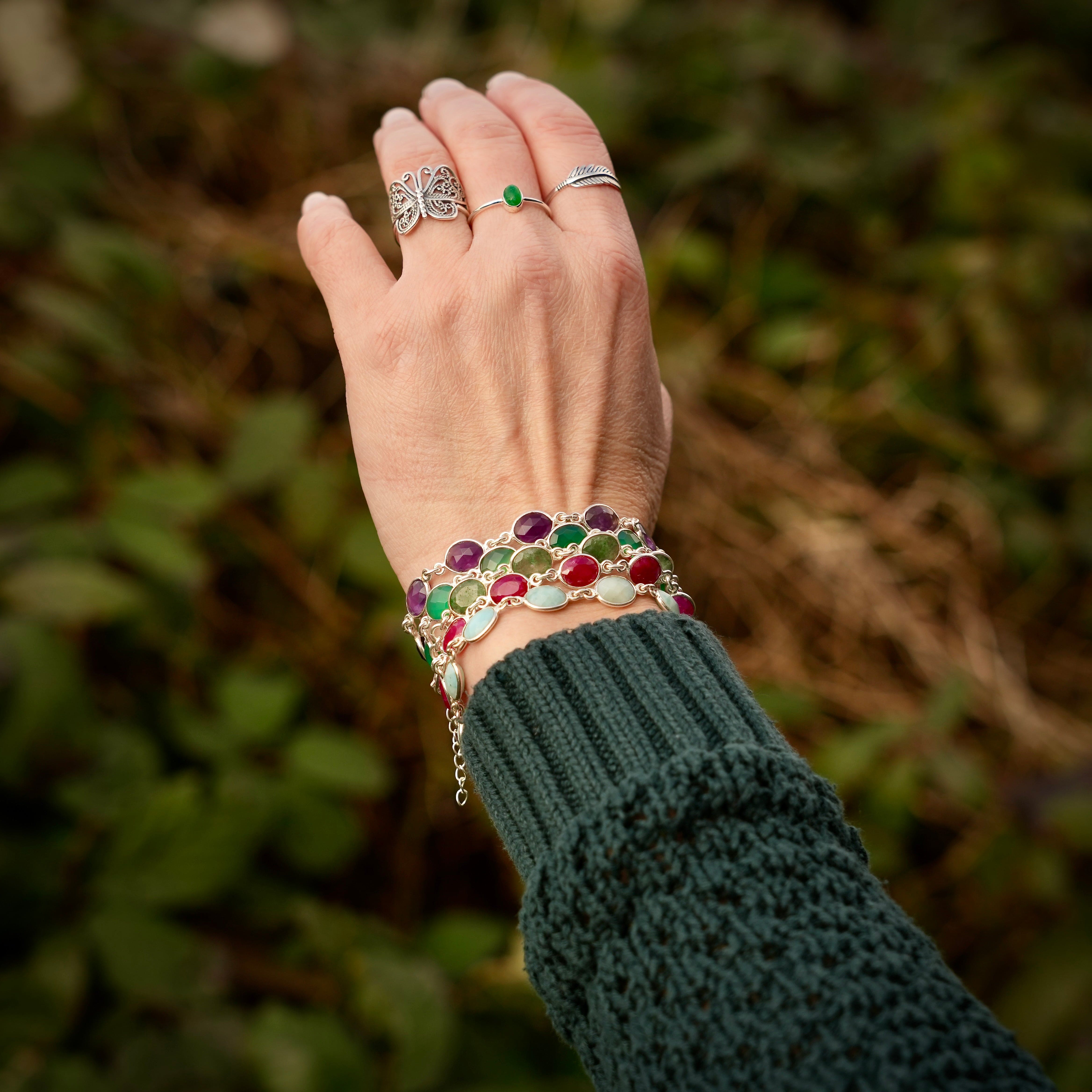 Hand met zilveren ringen, waaronder een ring in de vorm van een vlinder, een Zilveren minimalistische ring met een ovale Aventurijn edelsteen, en een ring in de vorm van een veer. Om de pols zitten armbanden met verschillende edelstenen.