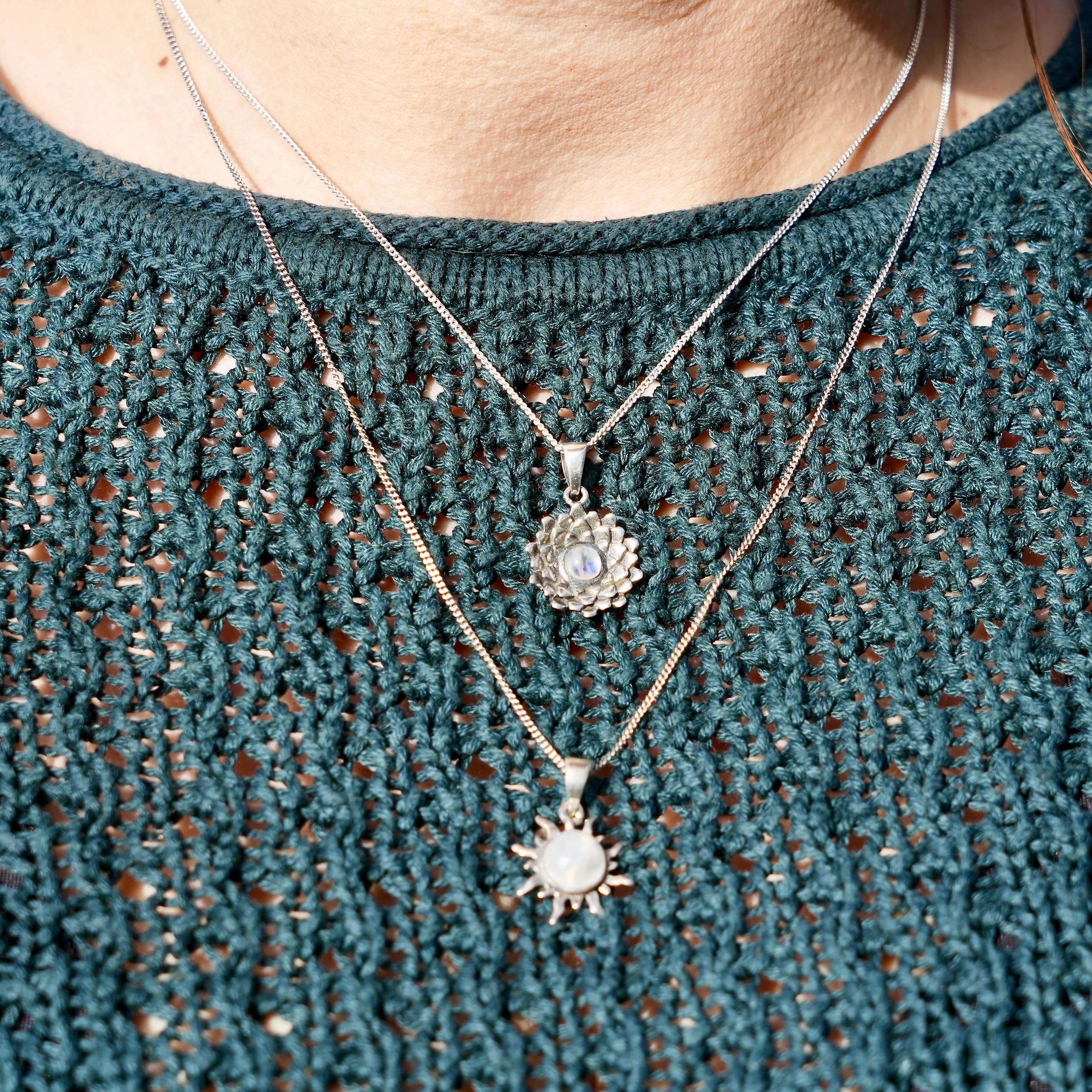 Een persoon met groene trui en twee halskettingen met een hanger in de vorm van een zon met maansteen en een hanger in de vorm van een bovenaanzicht van een lotus met een regenboog maansteen in het midden.