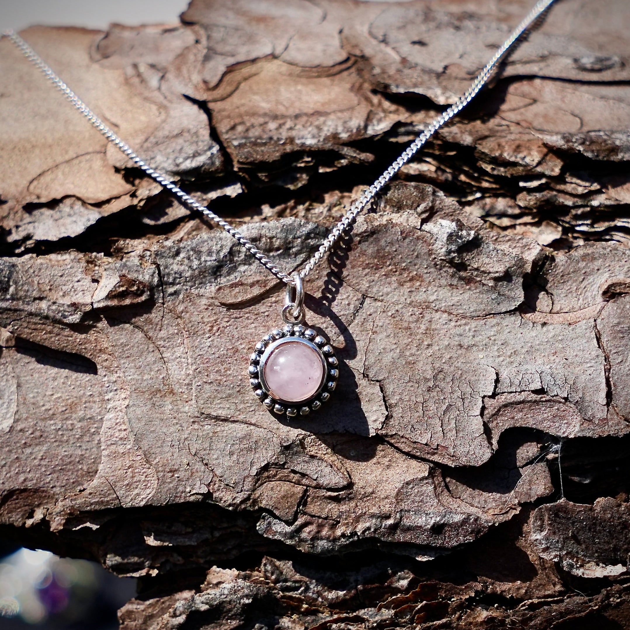 ronde zilveren hanger met rozenkwarts edelsteen en afgewerkt met zilveren stippen op een dennenboomschors