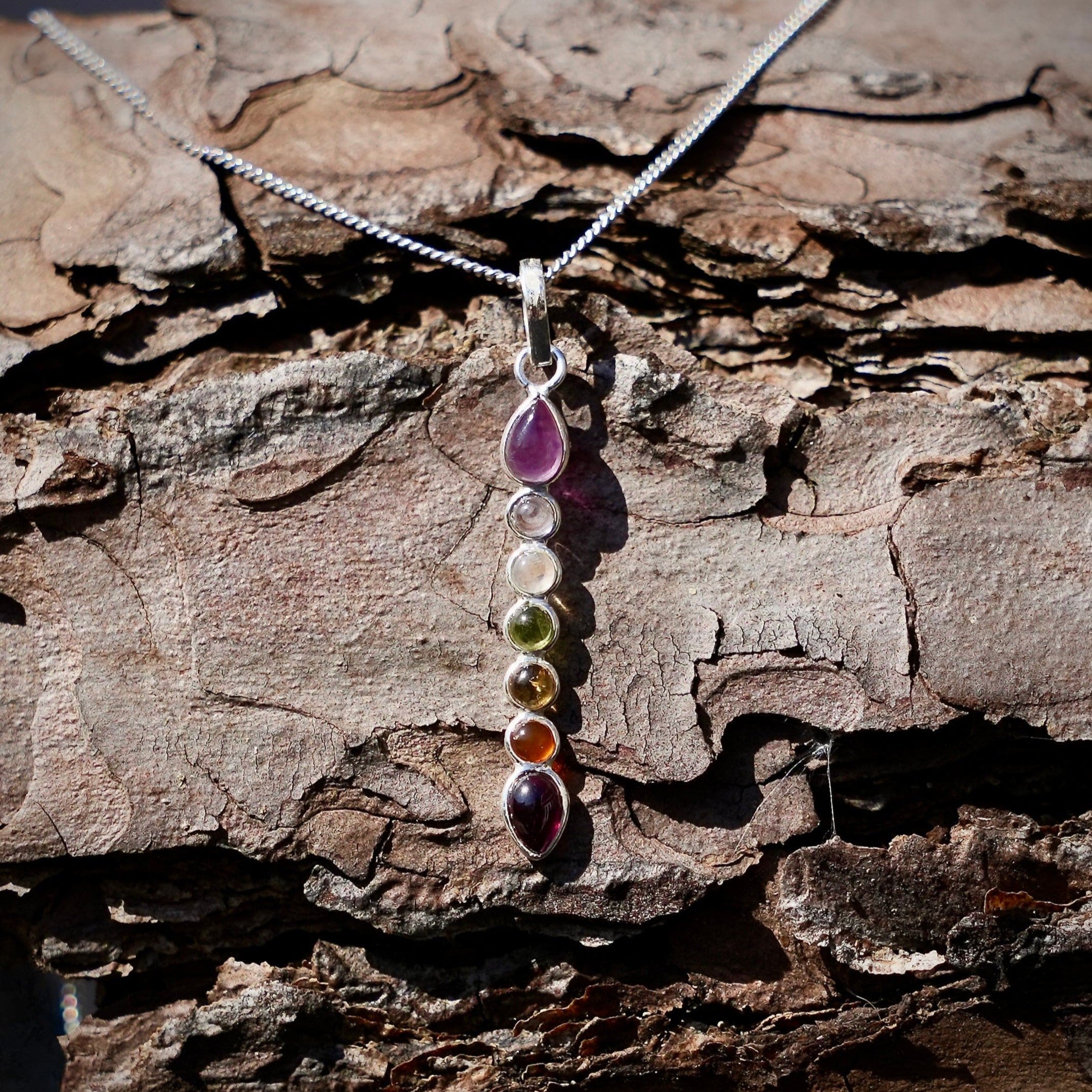 Chakra hanger van zilver met verticaal chakra edelstenen aan een halsketting over een stuk schors van een den